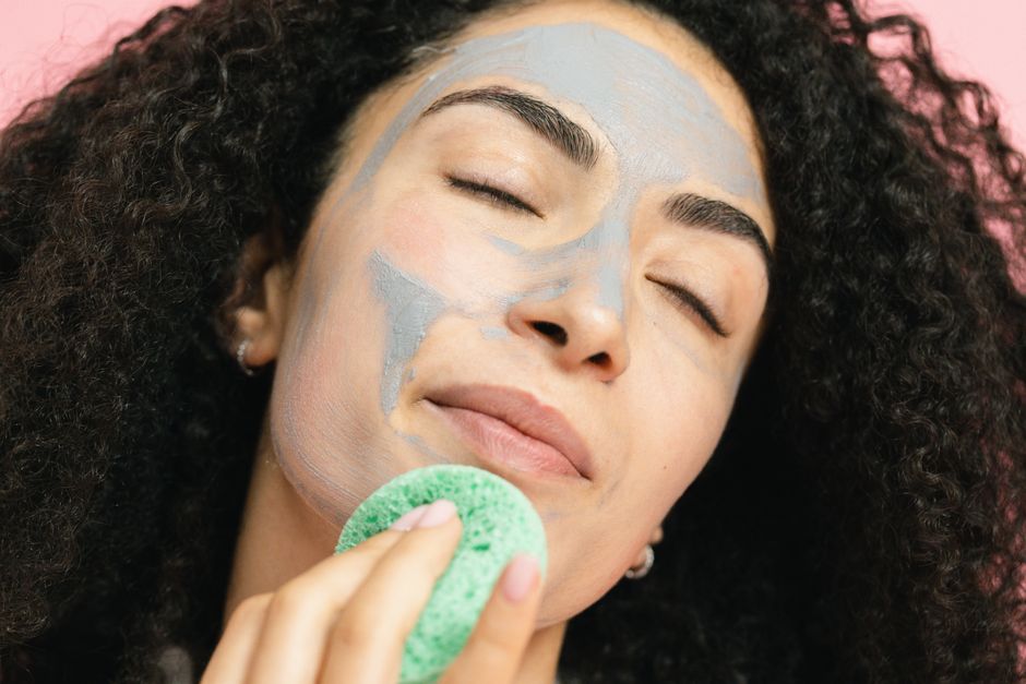 A woman with curly hair enjoys a relaxing facial treatment with a clay mask and sponge.