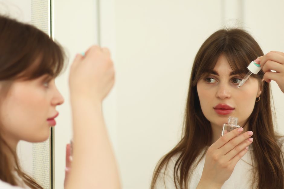 A young woman applying skincare serum in front of a mirror indoors.