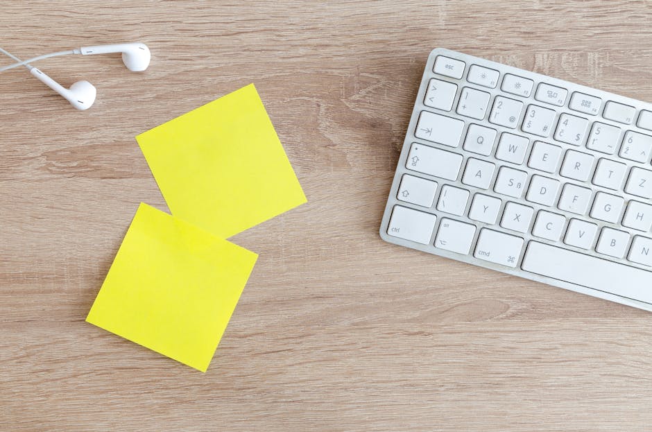 Top view of a workspace with a keyboard, earphones, and yellow sticky notes on a wooden desk.