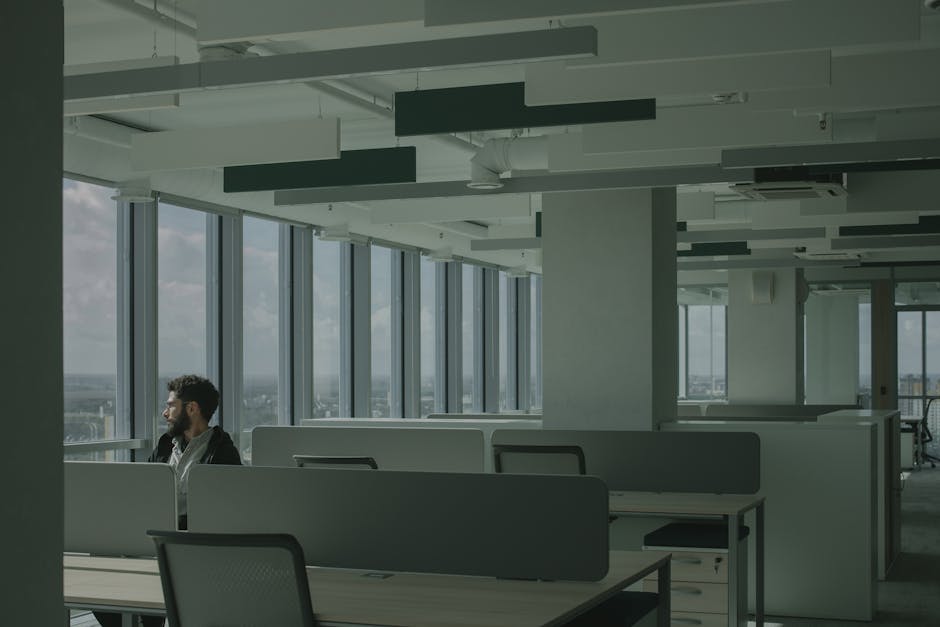 A man sits alone in a modern office space with large windows overlooking a cityscape.